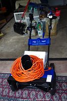 Front view of Snow Joe electric snow thrower with orange extension cord on top, manual visible behind, showing blue and black body and controls.