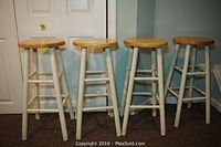 Four wooden bar stools with round natural wood tops and white legs, positioned side by side against white doors, partially on carpet.