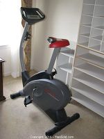 Full view of Pro-Form GT85X stationary exercise bike showing gray and black body with red seat, set up indoors near window and shelves.