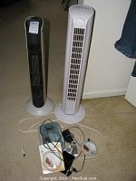 Two tower fans, one black and beige Duracraft, one white Ionic Breeze, with assorted cables and devices placed on floor in front.