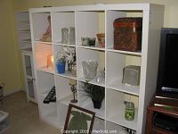 Off-white laminate shelf unit showing nine cubbies filled with various household decorative items in a living room setting.