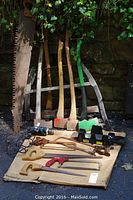 Wide view showing all items including axes leaning against wall, assorted saws, black saw blade stands, and electric drill.