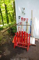 Lot contents showing snow shovels, brooms, red wooden sled, and primitive tiller against white wall.