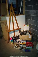 Wooden easel standing with two canvases, assorted airbrush boxes, manuals and tools on the floor and in a box next to the easel.