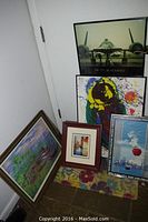Five framed prints grouped on floor in corner against walls, showing variety of styles and frame colors.