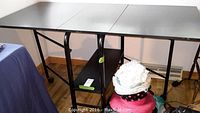Front view of the black folding table fully extended showing metal legs, plywood top, and lower shelf.
