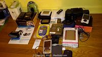 Wide shot showing all electronic items including boxed iPod, Kodak video camera, Vuescape camcorder, Zune with accessories, RCA CD player, Sharp calculator, remotes, and cables.