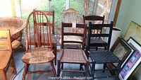 Seven wooden chairs of various styles arranged indoors near sliding glass doors. Some chairs have woven rush seats, others have wooden or upholstered seats.