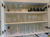 Full view of cabinet shelves containing various sets of glassware including goblets, tumblers, mugs, creamer, sugar bowl, and glass bowls.