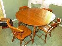 Round maple finish dining table with four matching chairs shown in room on carpet, table with turned legs and rounded corners.