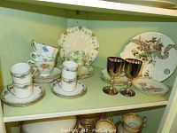 Shelf display showing multiple tea cups and saucers with floral decorations and gold trims, plus two reflective metal goblets and several decorative plates in the background.