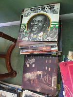Stack of vinyl record albums including Louis Armstrong and Neil Sedaka titles on a carpeted floor, beside a wooden chair leg.