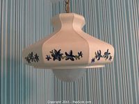 Front view of the ceramic hanging lamp showing the hand-painted blue floral design on a white cracked shade against a striped bluish wallpaper background.