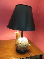 Full view of folk art style table lamp on wooden surface with black shade and bird-shaped pottery base.