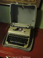 Vintage Olympia typewriter inside an open grey fabric-lined hard carry case on a wooden surface.