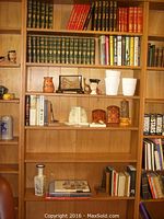Wide view of wooden bookshelves filled with multiple hardcover books and several decorative items including owl figurine, framed artwork, stone bird bookends, pottery, white ceramic pots, and candles.