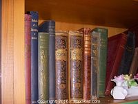 Shelf view of several antiquarian books with ornate covers and decorative binding, next to two small white ceramic floral decorations.