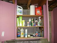 Wide view of cleaning supplies on wooden shelves inside a pink painted cabinet, showing multiple containers, spray bottles, a paper towel roll, and cleaning products.