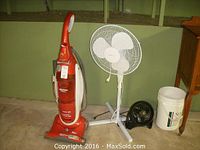 Wide view of orange Eureka upright vacuum cleaner, white pedestal fan, black table fan, and adjacent white bucket.