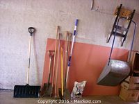 Garden tools including shovels, rakes, pole, and mounted workbench with metal vice, leaning against wall.