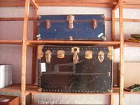 Two vintage trunks stacked on wooden shelving; one black trunk below and one blue trunk above, showing metal hardware and wear marks.