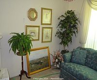 Wall with four framed gold-toned art pieces and one small framed mirror. One large framed print leaning against wall. Large faux leafy plant next to window. Floral arrangement near floor beside sofa. Blue floral patterned sofa partially visible. Wooden pedestal plant table holding a faux fern.