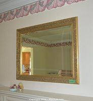 Large ornate gold framed wall mirror mounted on wall above white furniture, showing reflection of room.