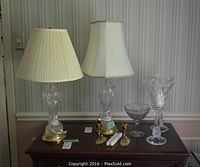 Two crystal table lamps with cream pleated shades on a wooden table, two lead crystal candle holders, two brass wall candle sconces, and a lead crystal candy bowl arranged next to lamps.