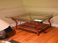End table with beveled glass top, metal frame, and rattan bottom shelf shown from an elevated front-left angle on hardwood floor.