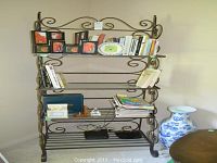 Full view of metal bakers rack with four glass shelves, holding various books and decorative items plus vase placed on floor beside rack.