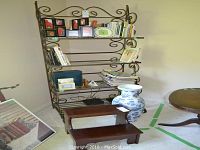 Wide view showing the metal shelving unit with wooden shelves containing books, framed pictures, board games, a large blue and white ceramic vase, and a wood two-step step stool in front.