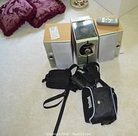 RCA RS2043 sound system with two speakers, remote control on top, black Minolta camera in carrying case, and Bushnell binoculars with carrying case in front