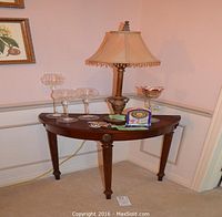 Photo of a half moon wooden table with carved legs placed against a wall. On the table sits a lamp, three glass candleholders, a small clock, and glass decor.