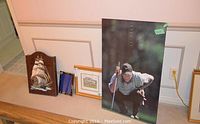 Wide shot of the dartboard case, golf prints, and decorative ship plaque leaning against a wall.