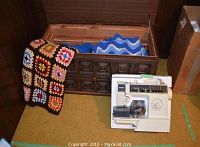 Full view of vintage wooden blanket chest open showing blankets and crocheted covers inside, with Singer sewing machine beside it on floor.