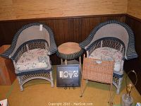 Two resin wicker armchairs with floral cushions, round resin wicker table, wicker magazine rack, framed LOVE artwork, and glass lamp in a corner room.