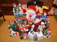 Wide shot of assorted Christmas decorations including stuffed snowmen, Santa figurines, wreaths, ornaments, nutcracker, snow globes, and lights arranged on a carpet near a fireplace.