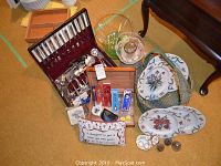 Photo showing wooden chest with silver-plated flatware including Oneida 1881 Rogers, glass dishes, collector spoons, decorative plates with floral designs, and small silver-plated items on carpet.