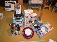 Full lot overview showing Cuisinart popcorn maker, Nutri Ninja blender with jar, Bissell spot lifter, T-Fal dessert plate, Starfrit apple peeler and stainless steel kettle on boxes and carpet.