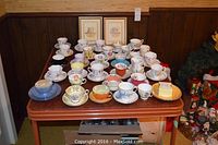 Tabletop overview showing over twenty bone china teacup and saucer pairs, two framed prints on wall, and hat box storage case partially visible.