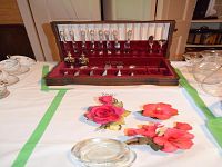 Closed wooden flatware chest with red velvet interior, flatware visible inside, Capodimonte floral decor and glassware on table around it.