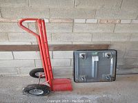 Side and angled view of red metal moving dolly and gray flat moving trolley against a brick wall.