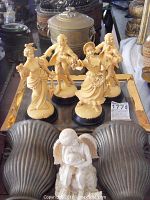 Four plastic Victorian style dancing figurines on black bases displayed on a glass tray with a bronze urn and other items in background