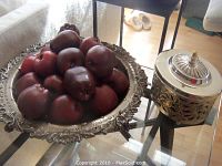 Photo showing large silver plated ornate footed bowl filled with dark red and red decorative apples on a glass table.