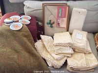 Photo showing folded Fingal Irish linen table linens in original boxes, various folded linens with beige and cream Damask patterns, and large woven throws with paisley patterns in red and green colors.