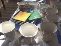 Photo showing eight McKenzie-Childs coffee mugs with pastel patterns surrounding stack of dinnerware plates and colorful leaf design coasters on top.