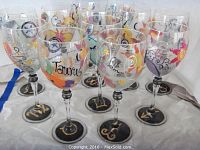 Set of twelve clear wine glasses with colorful hand-painted zodiac signs and symbols, shown on white cloth.