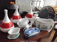 Overall lot on table showing three CB2 Paloma vases with white and red design, three black metal riveted baskets behind, and various ceramic decorative vessels including blue and white trinket box and cow creamer in front