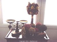 Brass decorative scale on wooden base surrounded by multiple decorative topiaries with dried botanical elements and berry clusters