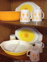 Shelf display showing 5 Fire King milk glass mugs with orange diamond pattern, 2 Pyrex divided dishes with yellow sunflower lids, and 1 orange Pyrex bowl.